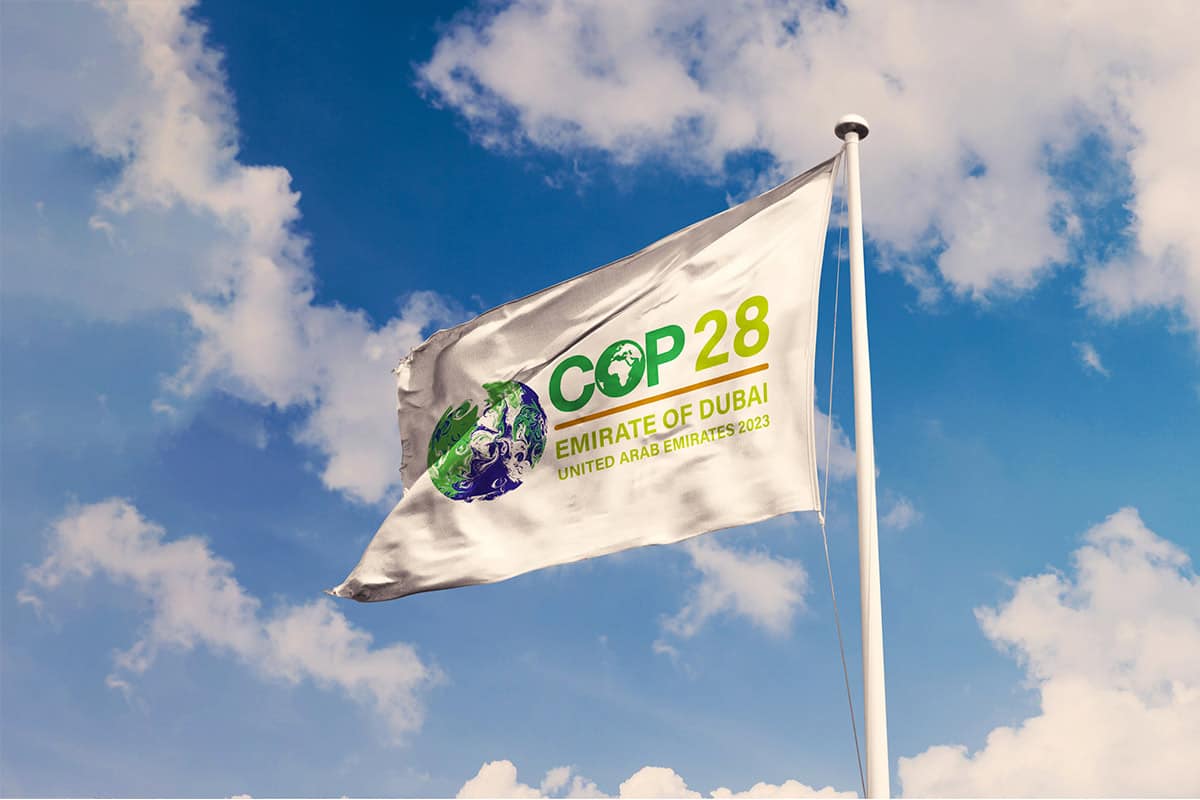 Eine weiße Fahne weht im Wind vor blauem Himmel und wenigen weißen Wolken. Auf der Fahne steht COP28, Emirate of Dubai. United Arab Emirates 2023. Links neben der Schrift ist eine stilisierte Erdkugel in grün-blau zu sehen.