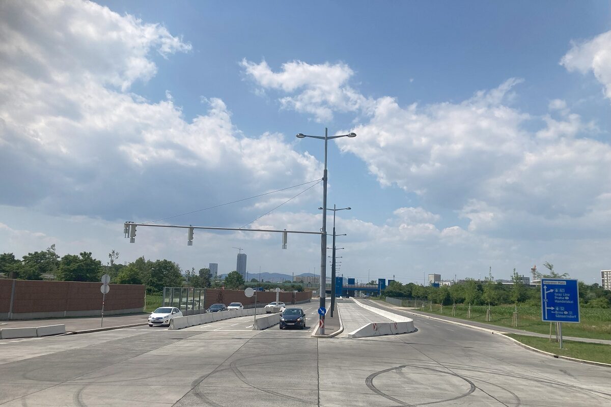 Breite asphaltierte Straße mit mehreren Fahrspuren, mittig durch Betonleitwände getrennt. Wenige Autos fahren bei Sonnenschein und leicht bewölktem Himmel in beide Richtungen. Links eine Backsteinmauer, rechts grüne Wiesen und ein blaues Verkehrsschild mit Richtungsangaben. Im Hintergrund sind vereinzelt Hochhäuser und ein entfernter Hügel zu sehen.