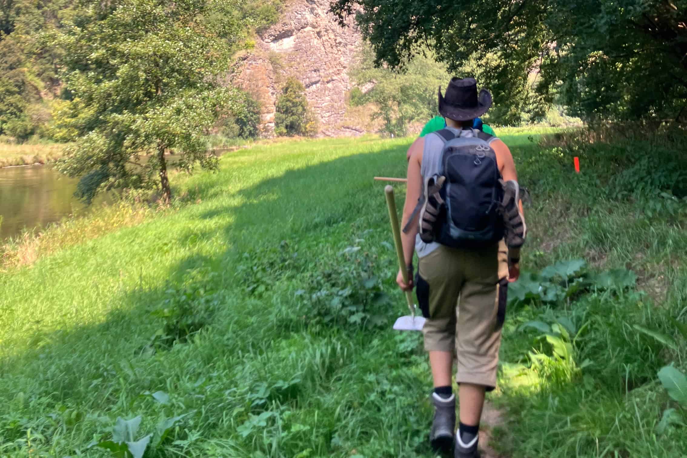 Eine Person auf einer Wanderung im Grünen. Man sieht ihren Rücken mit Rucksack. Links im Bild fließt ein Fluss.
